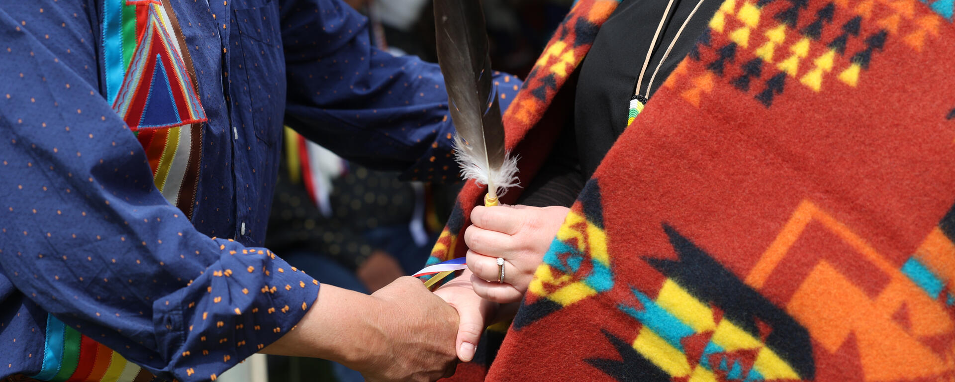 Someone in a ribbon shirt shakes hands with a person wearing a blanket and holding a feather.