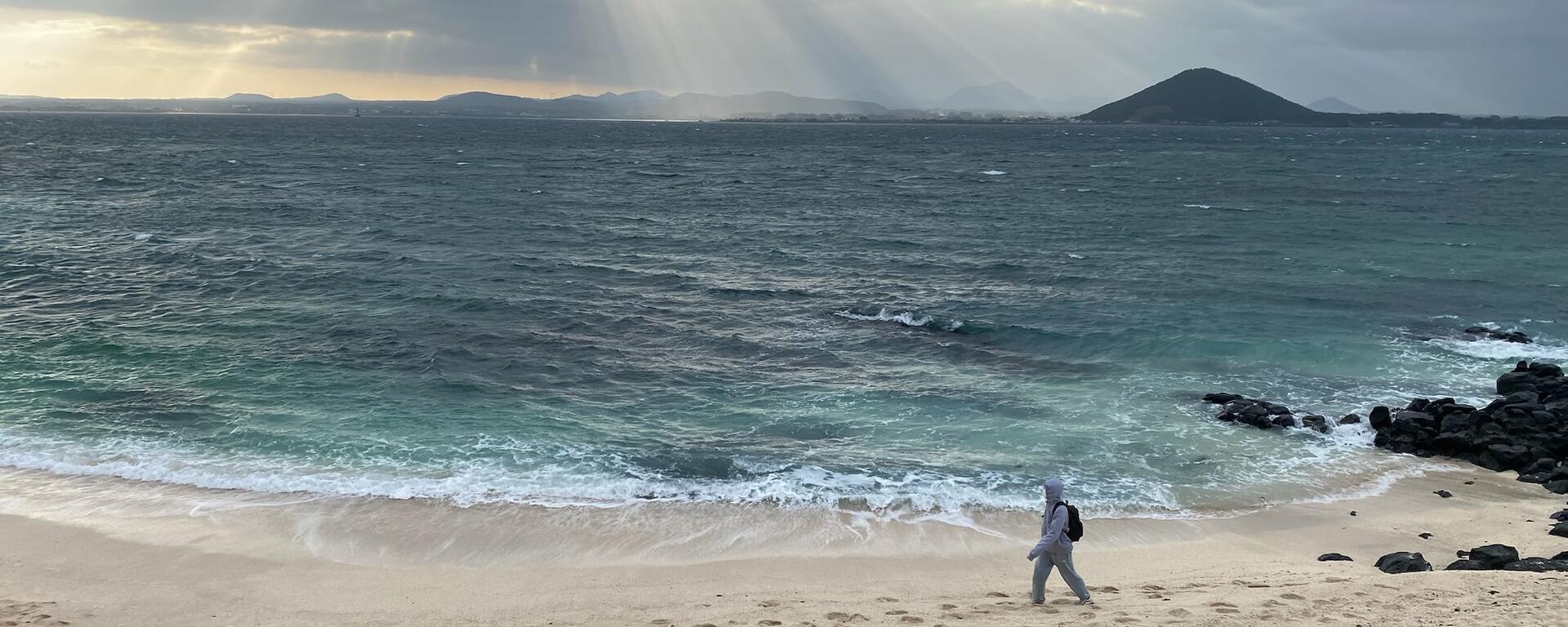 Looking down on a beach with stormy grey water. Sun rays illuminate the waves. A person walks through the sand. There is an island visible in the distant right.
