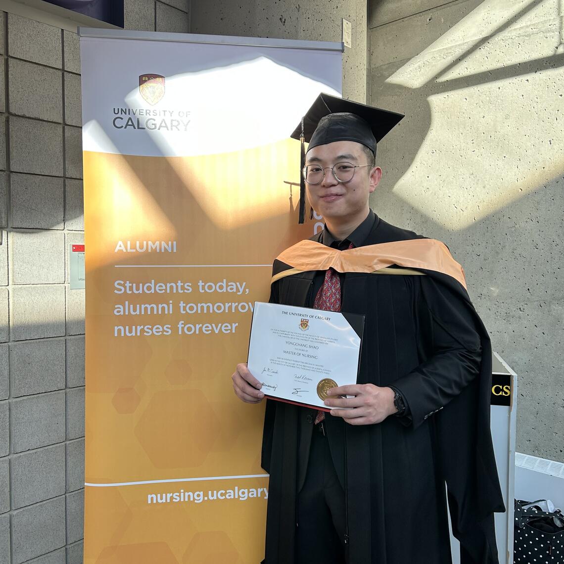 David Shao stands in front of a banner holding a diploma and smiling in graduation regalia