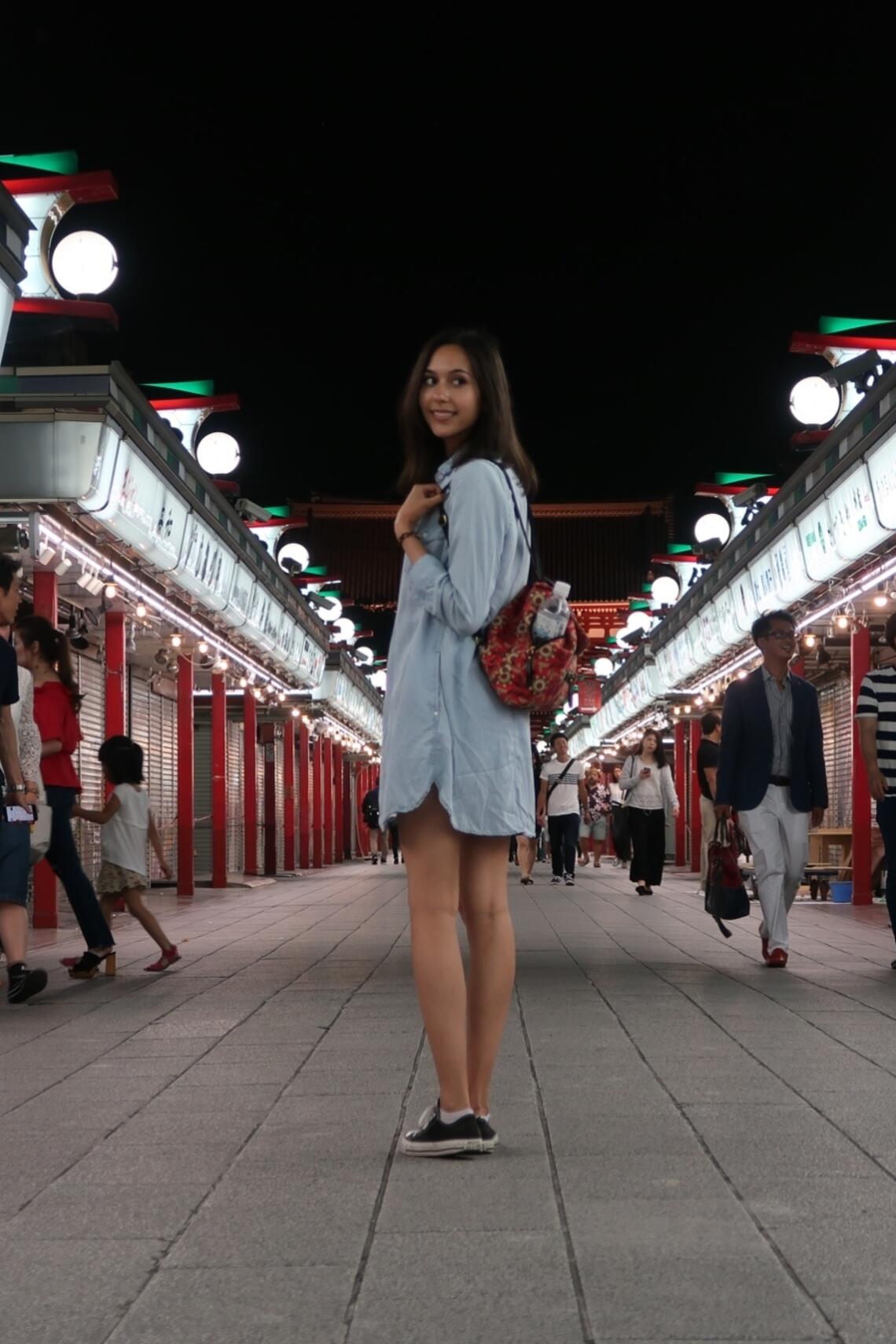 Young adult student with backpack turning towards the camera. They are in the middle of what seems to be a narrow East Asian shopping centre at night.