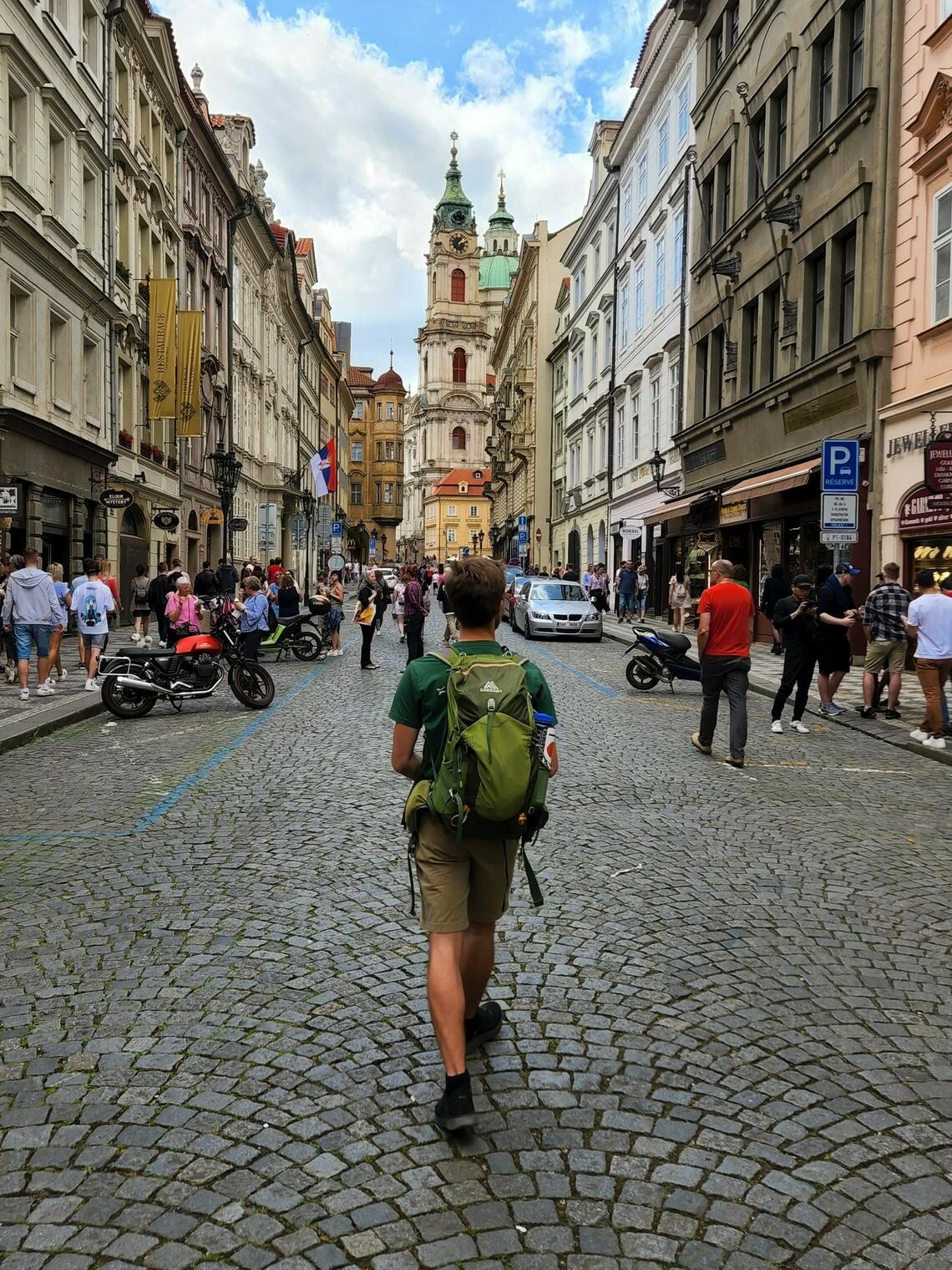 Image of a male presenting University of Calgary student with a green backpack walking away from the camera along a busy street in Prague Czechia.