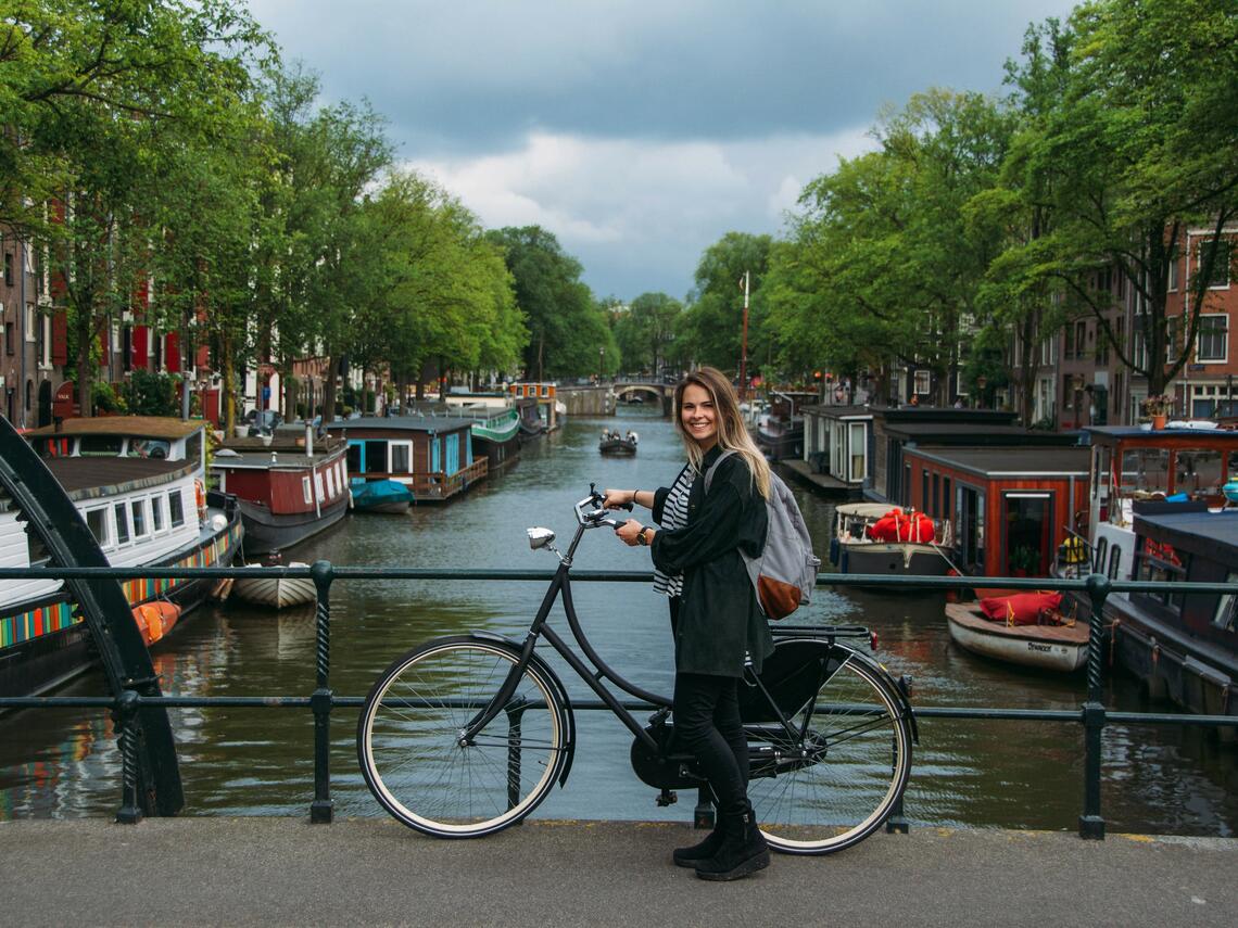 Image of a female presenting University of Calgary student in Amsterdam on a road bridge with their bike. Behind them is a lake filled with boats on the sides.