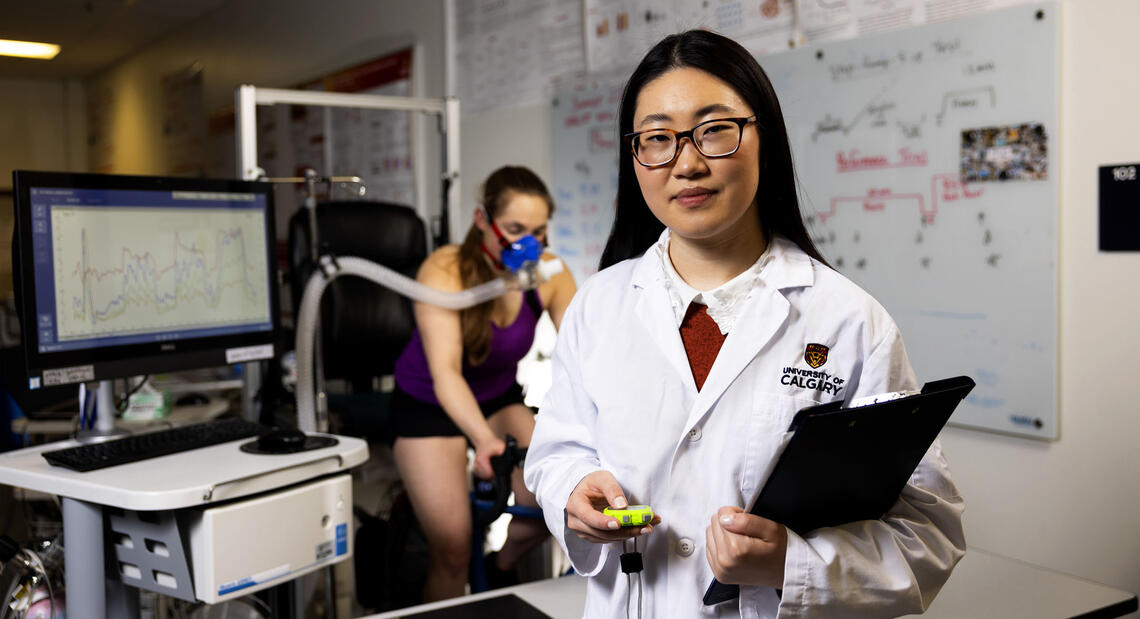 A woman in a white lab coat stands in front of a woman performing a test