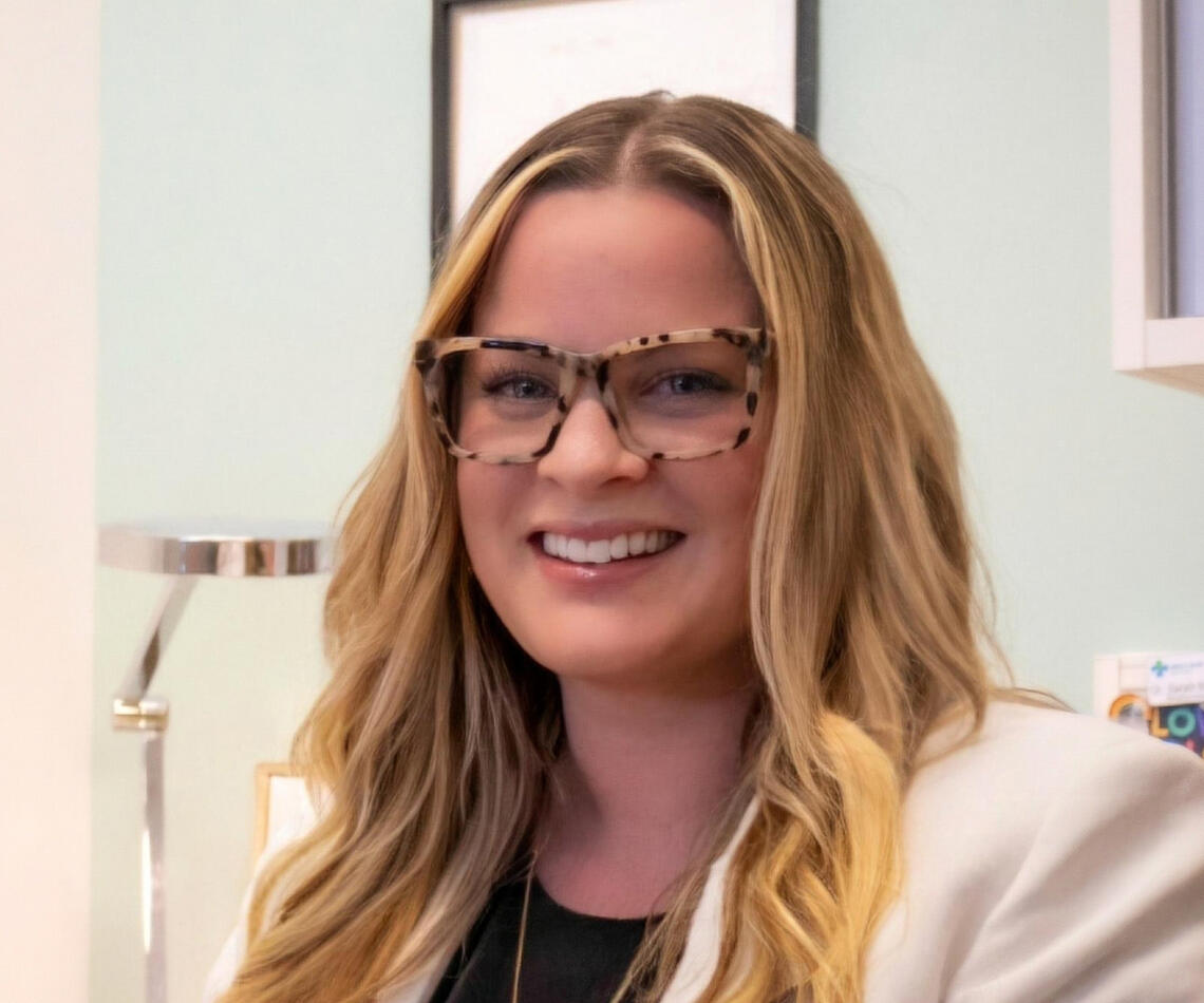 A woman in a white lab coat and glasses smiles at the camera