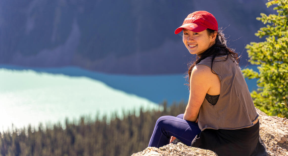 A woman in a baseball hat sits in front of a lake on a hike