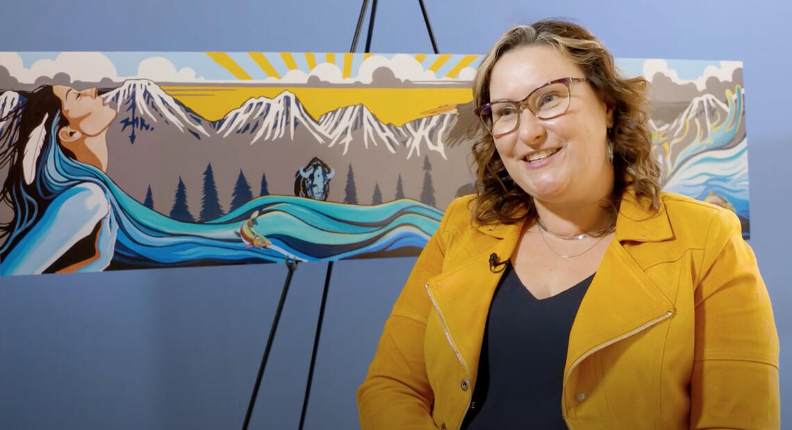 A woman sits in front of a painting of a mountain range