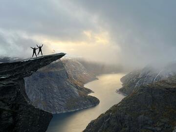 Two people jumping with hands up on a cliff overlooking cloudy fjords
