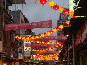 People walking in a street at dusk with red and yellow lanterns overhead