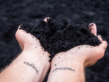 Photo taken looking down at hands clasping black sand; wrist tattoos read 'endless adventure'