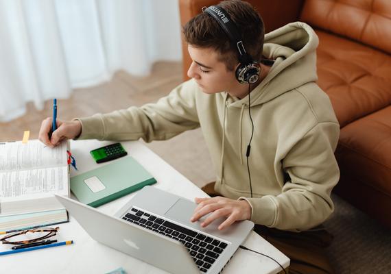 A young male student wearing headphones while writing notes. 