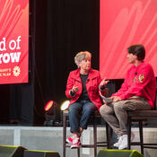 Two people in red talking on a stage with large screens behind them