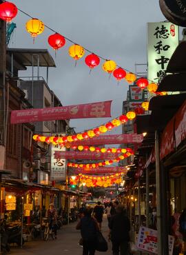 A dark street, lit by red and yellow lanterns strung overhead and neon shop signs. Two people walk ahead as silhouettes