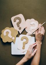 A variety of paper cut outs of question marks lying on top of a notebook, with a set of hands holding a pencil next to it.